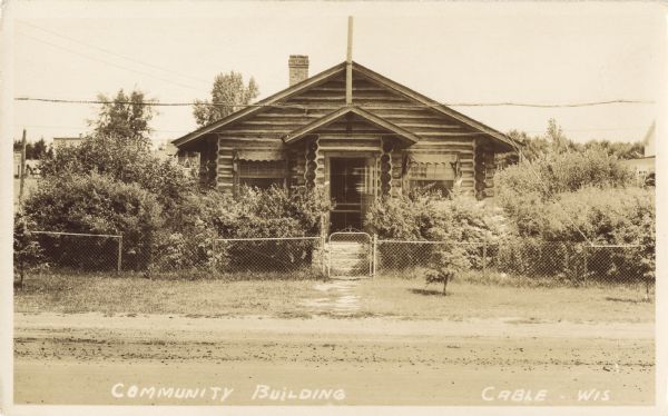 View from the dirt road of front of a one-story log building with a brick chimney. In the foreground are a chain link fence, gate, and shrubs. This building turned into the local Library.