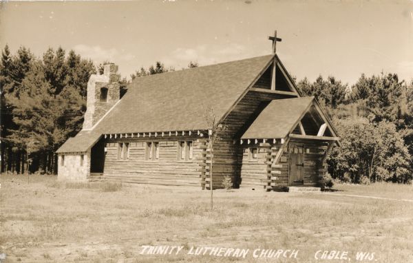 Exterior view of a log church showing the front doorway and entry way and the left side of the building. There is a stone chimney at the back near another entrance which has stone walls. There are trees in the background.