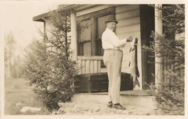 A man wearing a hat stands on the steps of the front porch of a cabin holding up a large fish at Cormican's on Clam Lake.