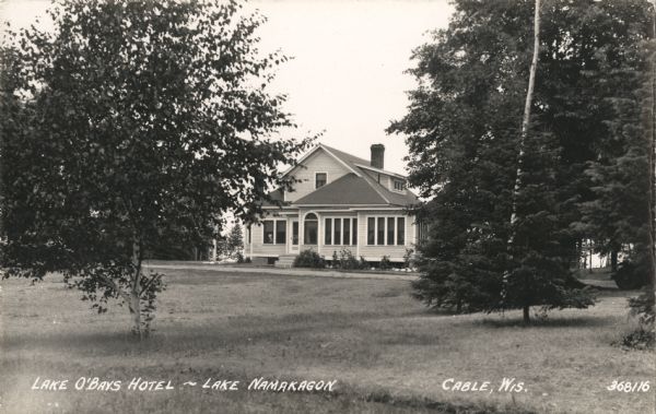 Exterior view across lawn of the Lake O'Bays Hotel on Lake Namakagon. The hotel is a two-story building with many windows. There is a road or path in front.