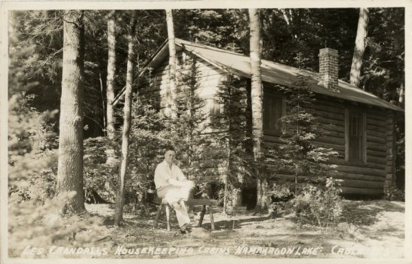 View of a man sitting on a wooden bench in front of small pine trees and a single-story log cabin with brick chimney.