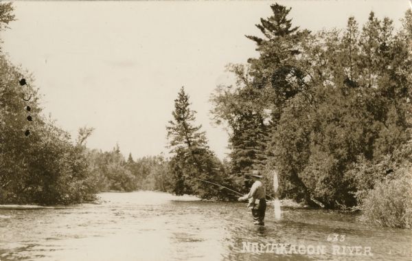View across water of a man fly fishing. He is standing in the Namakagon River in northwestern Wisconsin. Trees are along the shoreline on both sides of the river.