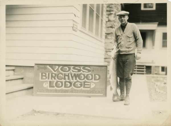 Outdoor portrait of a man wearing boots, a sweater and hat holding a large fish and standing next to a building and a sign that says "Voss Birchwood Lodge, Strictly Modern Accommodations." Voss Birchwood Lodge was on Spider Lake.
