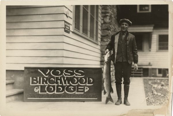 Outdoor portrait of a man wearing boots, a jacket and hat holding a large fish next to a building and sign that says "Voss Birchwood Lodge, Strictly Modern Accommodations." Voss Birchwood Lodge was on Spider Lake.