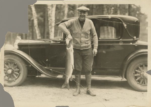 Outdoor portrait of a man wearing boots, pants, shirt, sweater and hat holding a large fish by its gills while standing in front of an automobile.