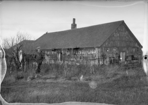 A man in work clothes and a hat stands in front of a weathered wooden building. A cat crouches near his feet, another sits to the right. Tall grass is growing around the building and trees are on the left. There are ghostly images of children moving near the building.