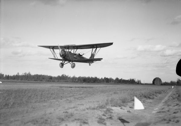 View of a man flying an airplane low over the airfield. He is wearing goggles and waving at the photogarpher. In the background is a barn and trees.