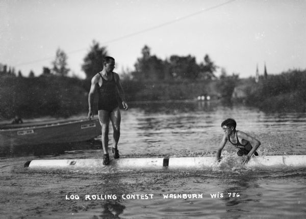 View from water of a log rolling contest. One man is still upright and the other has fallen into the water. A boat is visible to the left and trees line the shoreline in the background.
