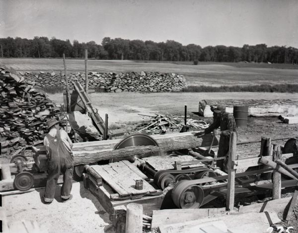 Elevated view of two men cutting logs at a sawmill. The men stand, one on either side, of a large circular saw, guiding a log through. Piles of cut logs sit to left and in the background next to a gravel road and an open field. Sawdust can be seen all over the working area. Trees are visible in the far background lining the field.