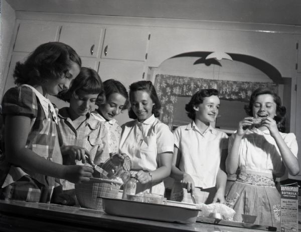 Six young girls in a home economics class stand around a table, measuring and pouring ingredients into a bowl. A girl to the far right holds a finished baked good in her hands, about to take a bite, while another girl, holding a knife, looks on. Flowered curtains can be seen above the window in the background. A package of Softasilk cake flour sits on the table to the right.