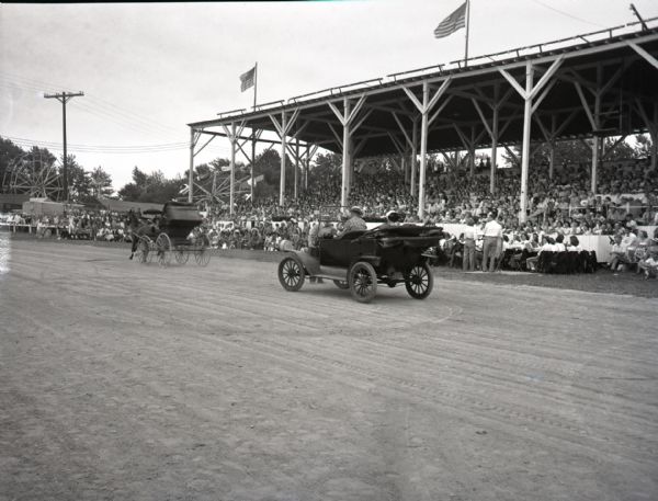 View from the inner track of a car and buggy race at a local fair. Crowds sit in the covered grandstand and a band sits at the ready on the sidelines. A Ferris Wheel and other carnival rides are visible in the background.