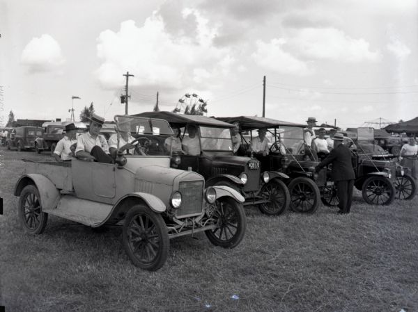 Men and women sit inside a row of four Ford Model T's in the parking lot at a fair in Wisconsin. Many of the men wear straw boater hats. Carnival rides, including a Ferris Wheel, and tents can be seen in the background.