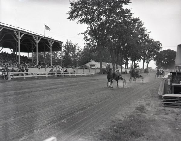 View from the inner track of harness racing at a local fair. The horses and their drivers can be seen racing around a tree-lined curve in the track and past the crowds in the grandstand to the left. An exhibition building is visible in the background.