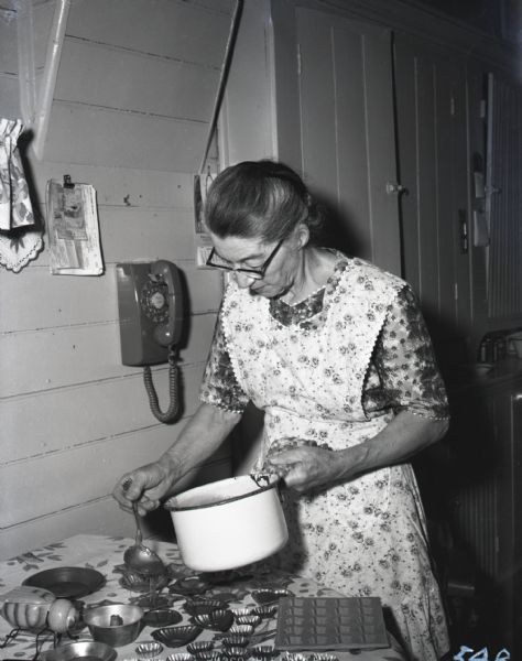 Mrs. Harvey Blue pouring maple sugar candies. Mrs. Blue, in a flowered apron, is holding a white enameled pot in one hand, and a ladle of maple sugar confection in the other, carefully pouring the mixture into molds of various shapes.
