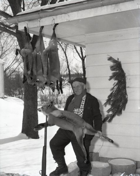 A hunter poses with his gun and a dead fox after a successful winter hunt. Three other foxes hang from the eaves of a house. Snow covers the ground behind him.