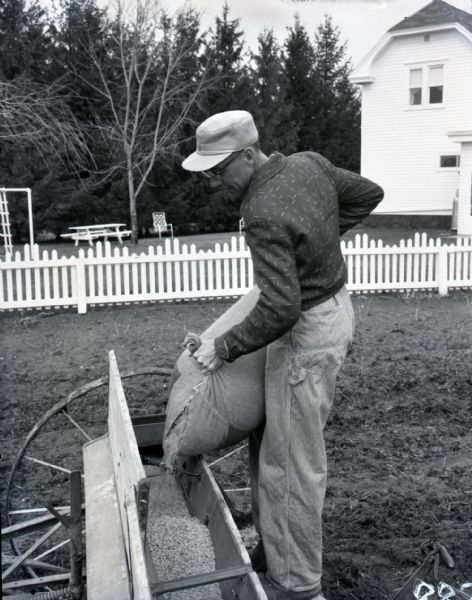 A farmer pours seeds from a canvas bag into the trough of a seed planting machine. A farmhouse, white picket fence, and picnic table with chairs are visible in the background.