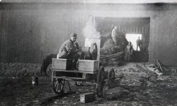 Three men from the Frank Otto threshing crew. Seated in foreground is Richarrd Krenke. In the background is Ed Fink of Antigo, and to his left is Bill Henchel of Oshkosh. Krenke sits on a wagon holding a steam engine while Fink and Henchel load hay into a threshing machine inside a barn.