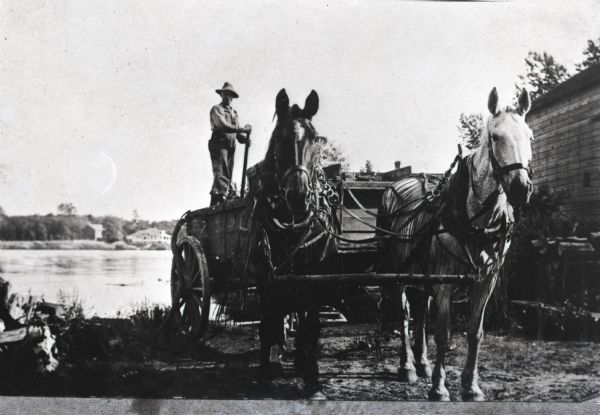 A man pumps water from the Wolf River into the back of a horse-drawn wagon to haul to a steam engine. There is a building on the left, and more buildings on the far shoreline.