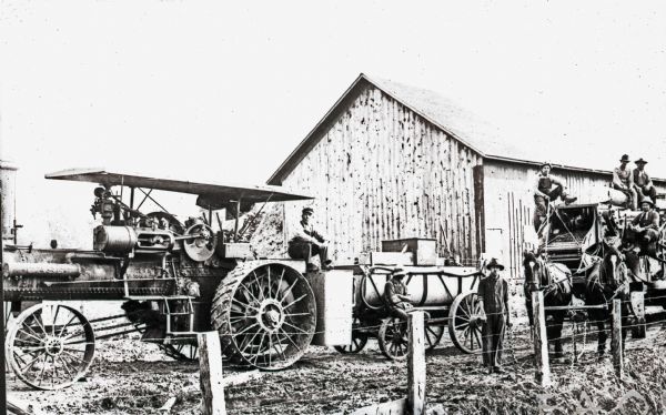 A threshing crew of men pose on tractors and threshing equipment on the Krenke farm. The tractor is powered by an engine and the threshing machine is pulled by a team of horses. A barn with what appears to be hay is visible behind them.
