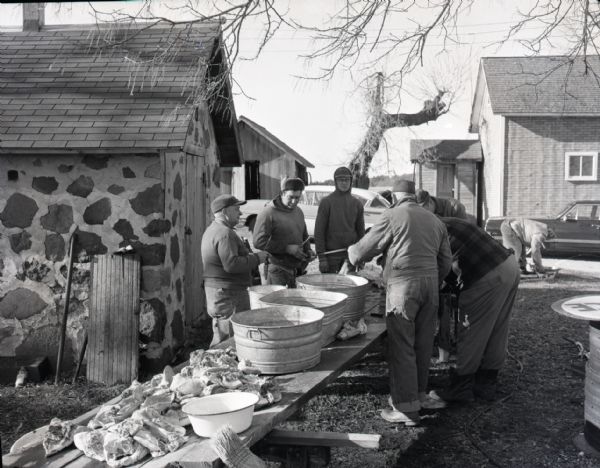 Six men of the Spiegel family stand outdoors around a long wooden table butchering chickens. The table is covered with large steel pots and pieces of meat. Another family member, cars, and the farmhouse are visible in the background.