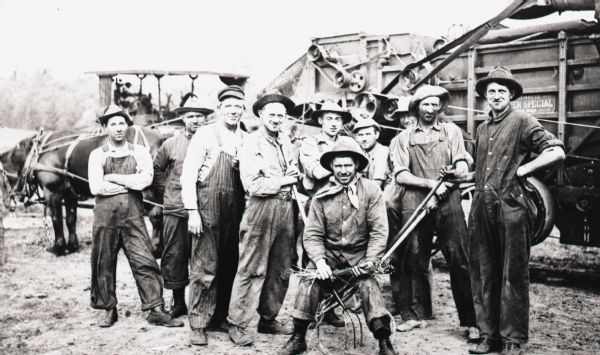 A group of farmer workers stand with pitch forks in front of the first gas engine for threshing on the August Krenke farm. A horse-drawn vehicle is also visible in the background.