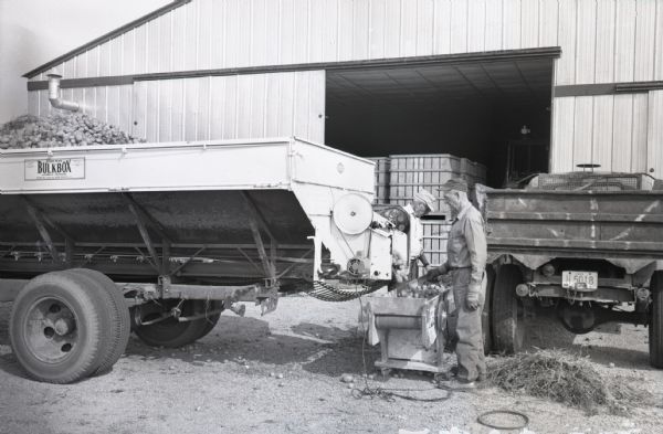 Potatoes going from truck to sorting machine at the Mumbrue farm. Roy Roth and George Mumbrue stand next to the machine in front of a barn.
