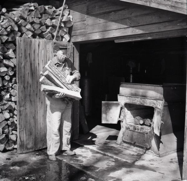 A man with an armful of wood gets ready to feed a woodburning stove inside a shed. A pile of cut wood sits behind the man.