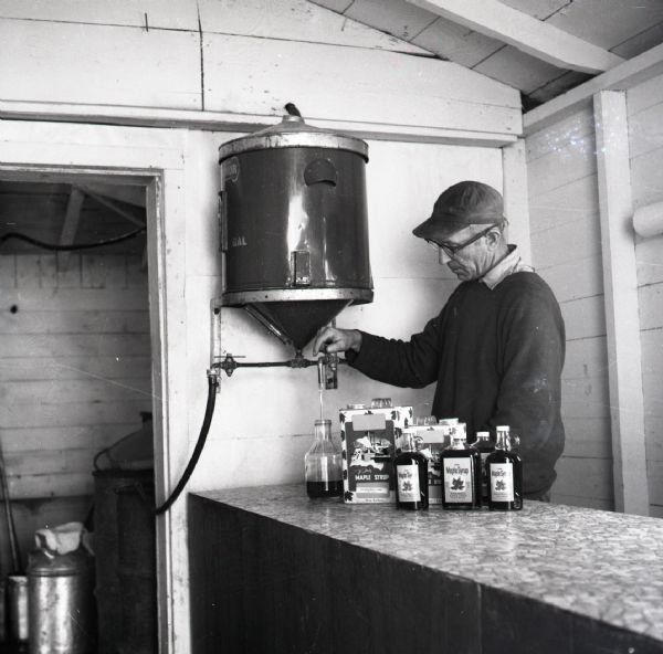 Milt Mehlberg filling bottles with finished maple syrup from his farm. Mehlberg is pouring syrup from a large sieve into a glass bottle. Several bottles and aluminum cans are on the counter.
