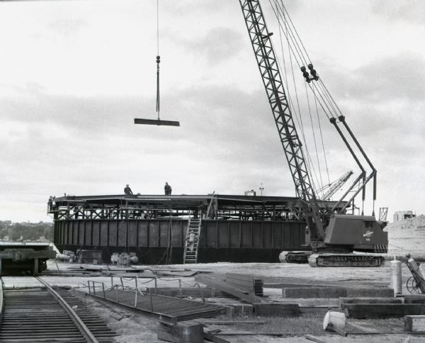 A crane lowers a beam onto the frame of a boat. Workers can be seen standing along the top of the vessel and scaling a ladder. Construction equipment and material litter the ground.