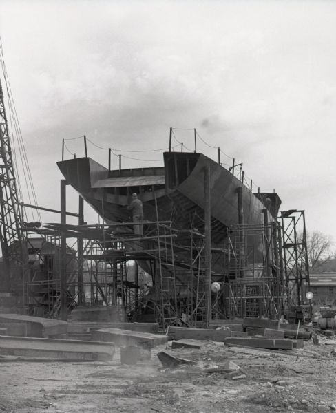 A man wearing a hardhat stands on scaffolding at the stern of a ship under construction. Beams and other shipbuilding material can be seen scattered around the construction site. A crane is visible on the left.