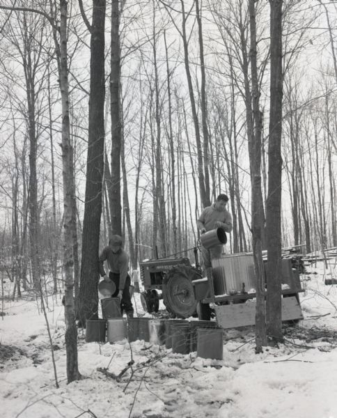 Two men in a forest are pouring maple sap into metal barrels and buckets at the Maple Crest Maple Syrup Farm. One man is standing in the back of the trailer hitched to a John Deere tractor, while the other man is standing nearby on the left. Snow is on the ground.