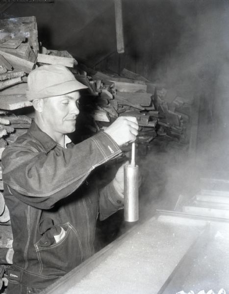 View of Wilmer Schulz, near High Cliff State Park, checking the syrup in a modern evaporator while standing over a vat of boiling syrup. Large piles of fuelwood are in the background.
