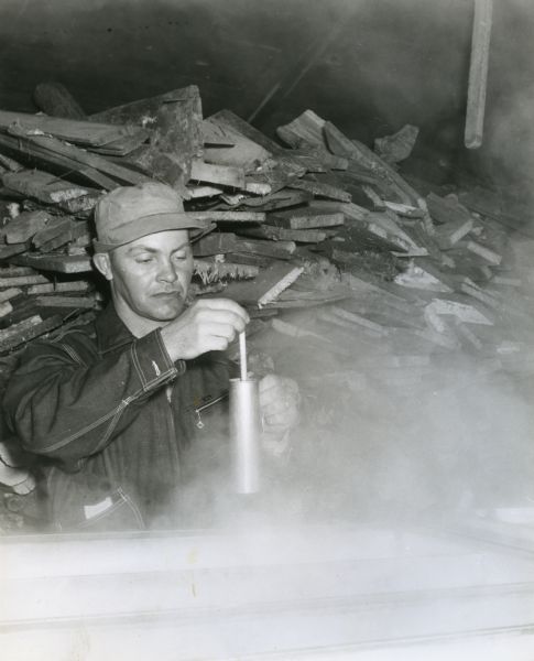 View of Wilmer Schulz checking the syrup in a modern evaporator near High Cliff State Park. Schulz is standing and holding a testing tool behind one of the boiling compartments of sap. A large pile of fuelwood is in the background.