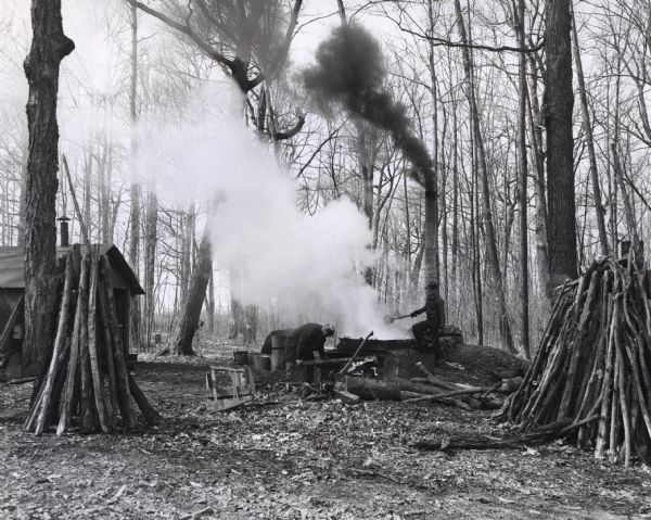 Two men are cooking maple sap over a wood burning cauldron in a clearing. One man is feeding the fire with logs while the other man is pouring in the sap. Smoke is billowing from the cauldron. A shack, and  logs stacked upright are in the foreground.