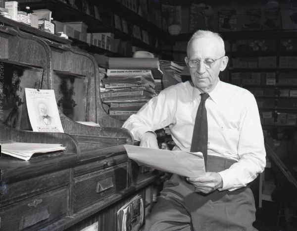 Howard Kimball is seated at his desk looking over papers in his Pine River general store. Various products are visible in the background, as well as stacks of John Deere manuals directly behind Kimball.