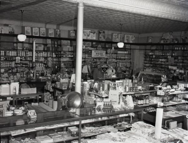 View of the interior of the Kimball Drug Store. A man and woman stand behind the counter in the very back of the store, partially obscured by a sign reading "Bakery Department." Linens, foodstuffs, and other household goods line the store shelves.