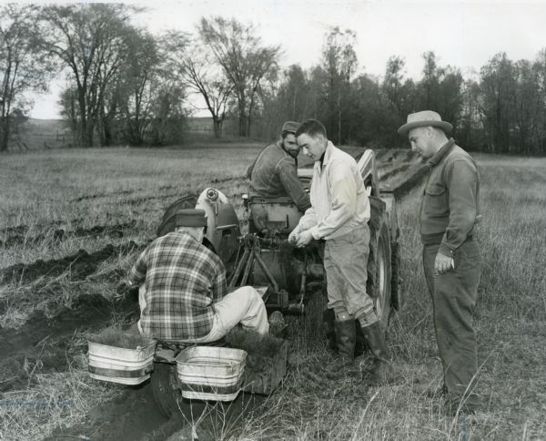 Four men prepare pine tree seedlings for planting in an open field. One man drives a small tractor, while two men stand behind. Another man sits on a tree planting trailer behind the tractor between two buckets filled with seedlings.