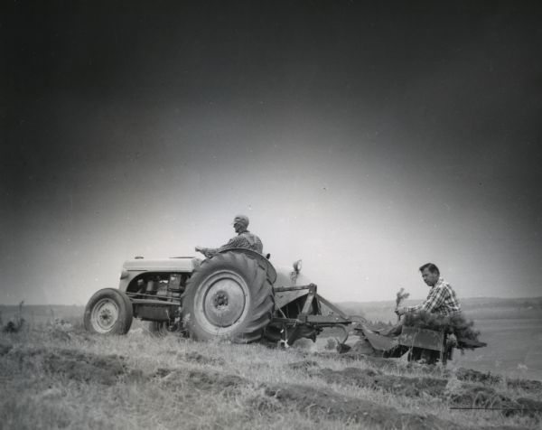Floyd Everett drives a tractor as Franklin Kempf plants Norway Pine seedlings using a tree planting trailer hitched to the back of the tractor.
