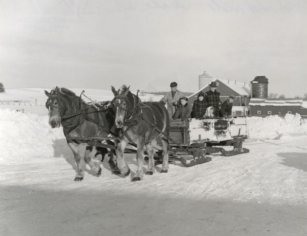 Winter scene with a farmer driving a team of horses pulling a large wooden sled on a snow-covered road. Standing to his side, from left to right, is Marge Mueller Van Heuklon and Mark Mueller, along with another man and two children. In the background are farm buildings.