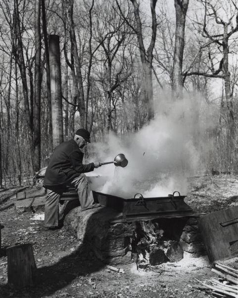 Harvey Blue, standing next to a wood burning fire pit. He is pouring maple sap into a large vat, which is in a forest clearing.