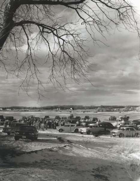 Shoreline view of people and cars gathered near a frozen lake. A few cars and ice fishing shelters are visible in the background.