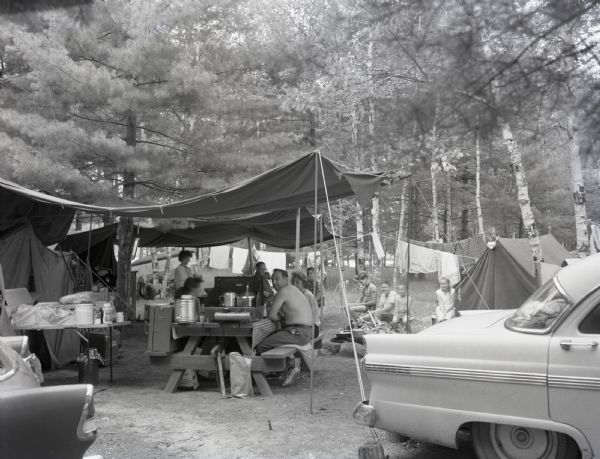 A campsite at Hiawatha National Forest. Men and women sits at a picnic table, while children sit by the campfire.