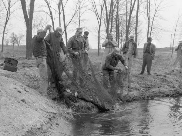 A group of men on a shoreline pulling a fish net from a lake. Several other men watch nearby. Two men use cameras to record the event.