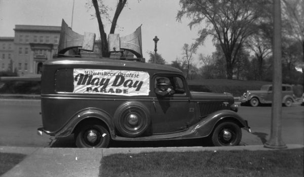 Side view of a car parked at curb decorated to participate in the May Day parade. The car has two large megaphones mounted on top and a banner that reads, "Milwaukee's Greatest May Day Parade." The car is parked on a quiet street and a man in a suit and hat sits in the passenger seat.