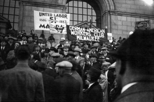 May Day rally with a crowd of Union members on the steps of a large stone building. Banners read "United Labor 51st May Day: 1886-1937"  "German-American Workers Club Milwaukee" and "United Trade Union."