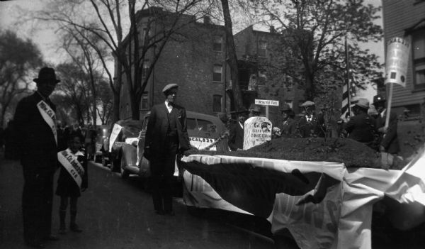 May Day parade with a closeup of a "memorial" float for company unions. A man leans on the parked float. The float features a faux grave and the gravestone reads "Gone but not forgotten." A banner on the side reads "Company Unions." A line of other cars in the parade stretches into the background. A man and a boy wearing Milwaukee Co. Industrial Union CIO stand in the foreground street. In the background are four-story brick apartment buildings and a small group of Union supporters one holding a sign.