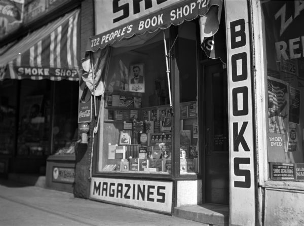 Storefront of the People's Bookstore showing a Lincoln and Jefferson display. An awning is over the adjacent smoke shop on the left, and on the exterior wall near the windows is a Coca-Cola sign over a peanut or candy dispenser. On the right is a storefront of a cobbler.