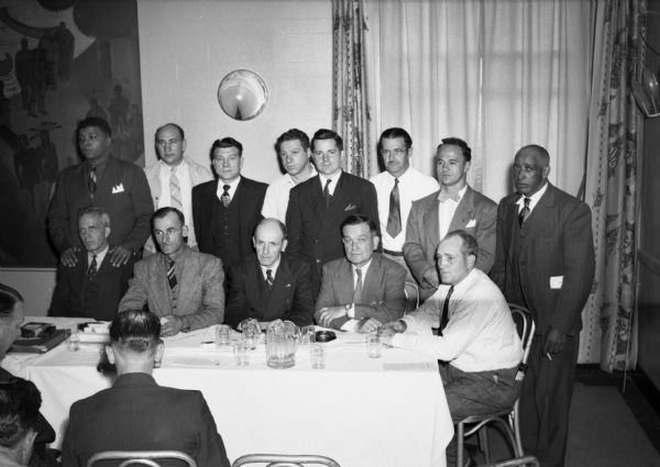 Posed group shot of the United Steelworkers of America District 32 Convention Resolutions Committee. The men wear suits and are all sitting or standing around a long table. On the table there is a pitcher, water glasses, papers and an ashtray. Hanging on the wall behind them are curtains, a painting of Native Americans and art deco light fixtures.