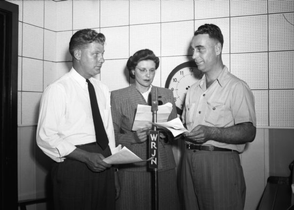 Pro price-control broadcast at WRJN. A woman and two men stand encircling a microphone holding scripts. A clock is on the wall behind them and a piano [?] is to their right in the small broadcast room.
