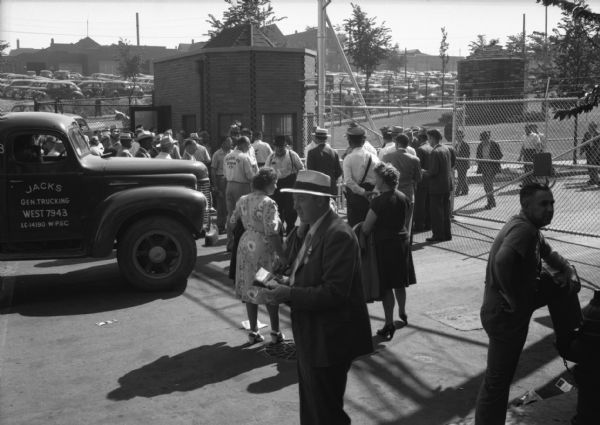 Members of the Farm Equipment Workers Union hand out leaflets at the International Harvester Co. plant during the Union's convention. A small crowd of people are gathered at the chain-linked gate of the factory. A pickup truck waits at the gate. Two brick guard houses are positioned near the gate and a large full parking can be seen in the background.